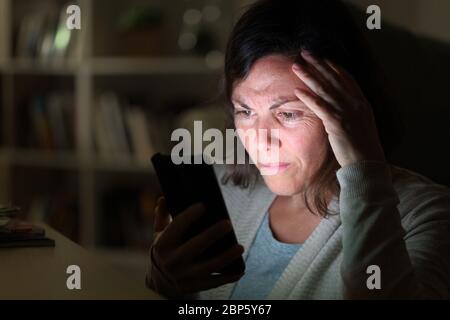 Preoccupied adult woman reading on smart phone lighted screen sitting at night at home Stock Photo