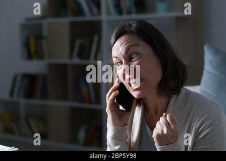 Angry adult woman calling on smart phone sitting in the living room at night at home Stock Photo