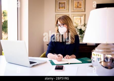 Portrait shot of middle aged woman wearing face mask for prevention while working at home on laptop during coronavirus pandemic. Home office. Stock Photo