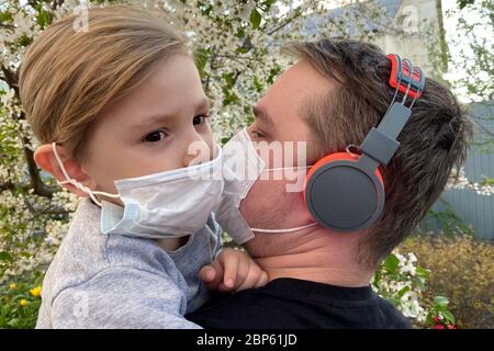 A little boy in medical mask hugs a man. Dad holds his son, they both are wearing the protective mask, quarantine virus protection concept. Stock Photo