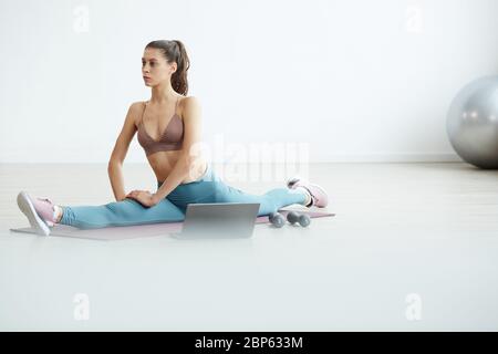 Minimal full length portrait of fit young woman doing splits during online workout via laptop at home, copy space Stock Photo