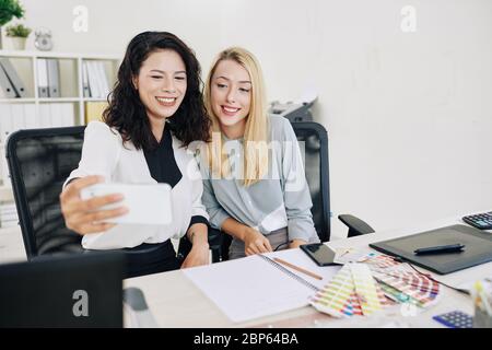 Cheerful young female designers sitting at office desk and taking selfie after work Stock Photo