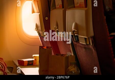 Food in brown paper bag put on plastic airplane tray table at seat back with blurred passenger hand opened black leather bag. Sunlight passing through Stock Photo