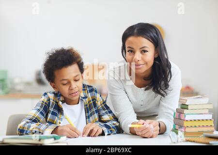 Portrait of young African-American woman smiling at camera while helping son studying at home, copy space