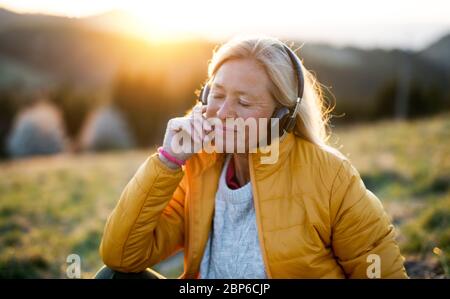 Senior woman sitting outdoors in nature at sunset, relaxing with headphones. Stock Photo