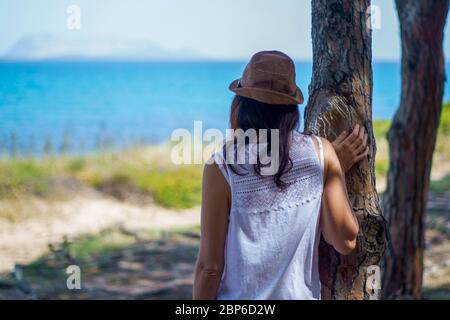 Pensive young woman with long hair and hat, seen from behind, looks at the sea leaning against a tree in a pine forest Stock Photo