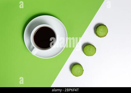 White cup of black coffee and three green french macarons on divided white and green background from above. Closeup of sweet breakfast, geometrical di Stock Photo