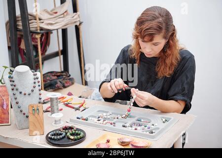 Young woman concentrated on making jewelry and using pliers to attach hook Stock Photo