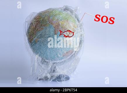 Text SOS and icon of fish on the Model planet Earth in polyethylene plastic disposable package on light grey background. Ecology problems. Concept pollution of environment with polyethylene plastic waste. World Earth Day. Save the planet Stock Photo