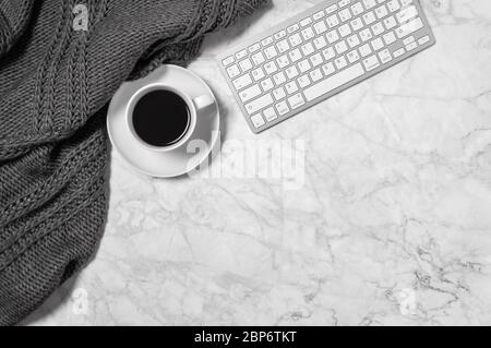 Marble desk with warm wool plaid or scarf, coffee cup and keyboard in black and white. Cozy home office workspace. Minimalist freelance or business co Stock Photo