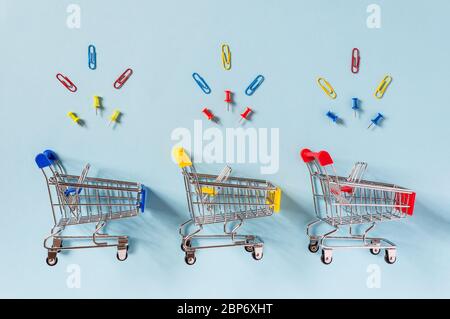 Colorful supermarket shopping trolley carts on blue background with yellow, red and blue clips and pins above. Shopping for office supplies, commerce Stock Photo