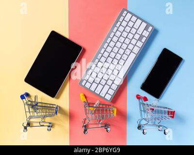 Online shopping concept with supermarket shopping trolley cart, mobile phone, tablet and keyboard on colorful yellow, red and blue background. E-comme Stock Photo