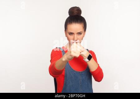 Let's fight! Portrait of determined aggressive confident girl with hair bun in denim overalls boxing with clenched fist, punching in front of camera, Stock Photo