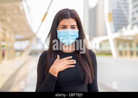 Sick young Indian woman with mask having sore throat in the city outdoors Stock Photo