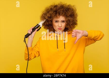 Upset displeased woman using curling iron to make afro hairdo and showing thumbs down dislike, ironing hair curls, expressing dissatisfaction with too Stock Photo