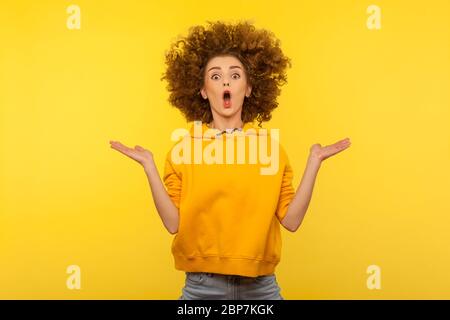 Portrait of extremely surprised girl in urban style hoodie jumping with her messy curly hair up raising hands and open mouth in amazement, shocked fac Stock Photo
