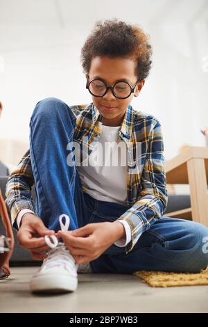 Full length portrait of cute African-American boy wearing glasses and tying shoes Stock Photo