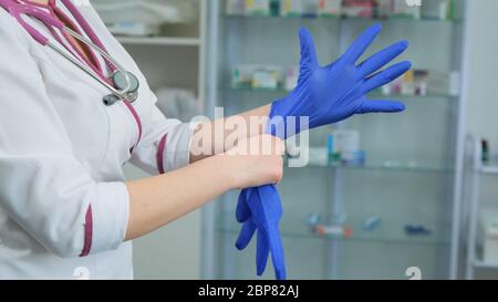 Young women doctor medic in white medical suit gown and violet stethoscope putting on rubber blue gloves on the left hand in hospital or clinic. Side Stock Photo