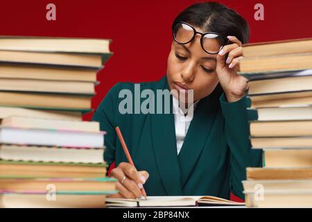 Portrait of tired female teacher sitting at desk between stacks of books and grading homework against red background Stock Photo