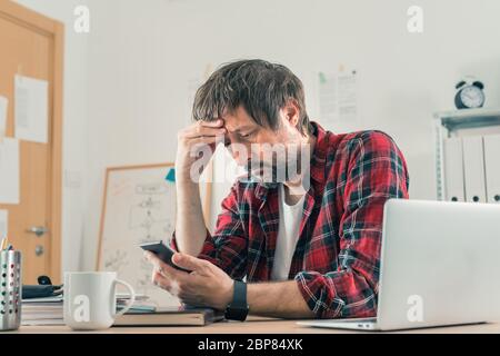 Troubled disappointed freelancer using smartphone for communication while sitting at the desk of his home office, selective focus Stock Photo