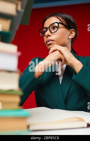Low angle view at young female teacher sitting at desk with stack of books in foreground, shot on red Stock Photo
