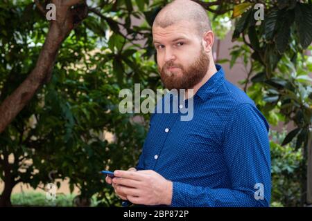 Bald bearded man in a blue shirt standing outside holding a smartphone Stock Photo