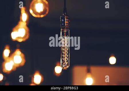 A thin long glass lamp shines in the dark against the background of other round incandescent lamps . Electricity. Lighting. Stock Photo