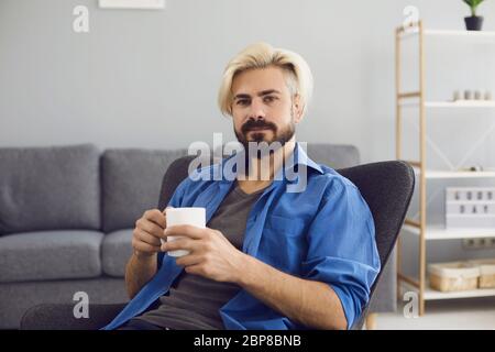 Online video chat. A young man with a mug of coffee looks at the camera in a living room. Stock Photo