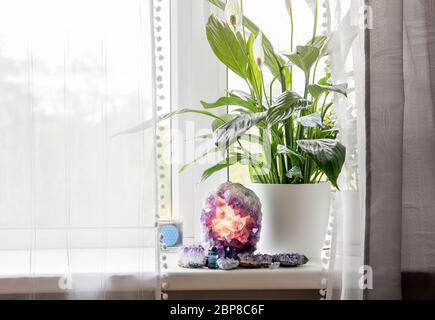 Using semi precious stone crystal details in home concept. Amethyst geode lamp burning, spiritual calming home atmosphere. Air plant Spathiphyllum. Stock Photo