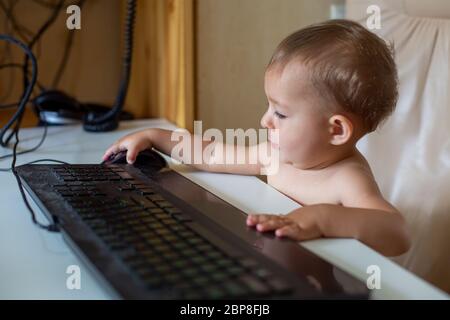 early study. cute little baby sitting on an office chair and before the black keyboard and puts hand on black mouse, he works, close up, soft focus. Stock Photo