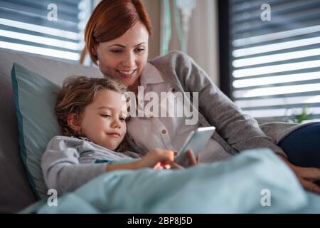 Small girl with mother in bed in hospital, using smartphone to pass time. Stock Photo