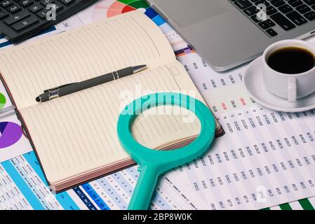 Desk of a businessman. A magnifying glass on a Notepad, a computer, a Cup of coffee, and tables with finances. Company documents and statistics Stock Photo
