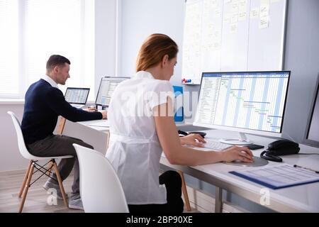 Businesswoman Analyzing Gantt Chart On Desktop In Office Sitting Behind Male Colleague Stock Photo