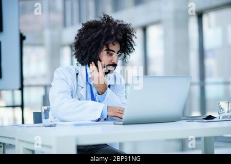 Portrait of male doctor sitting in hospital, using laptop and smartphone. Stock Photo