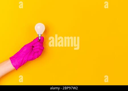 Hand in pink protective rubber glove with lightbulb isolated on yellow studio background with copyspace. Gesturing, holding, presenting things. Negative space for your advertising. Showing, pointing. Stock Photo