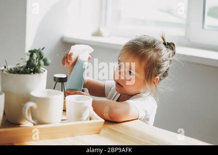 Funny cute toddler girl watering house plant on table in bright interior home Stock Photo