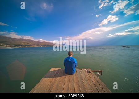 Male traveller sitting on a wooden pier and admiring the beauty of Ohrid Lake, Northern Macedonia Stock Photo