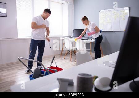Two Smiling Young Janitor Cleaning The Desk And Mopping Floor In The Office Stock Photo