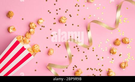 Popcorn in red and white paper bucket scattered on pink background with golden ribbon and confetti. Stock Photo