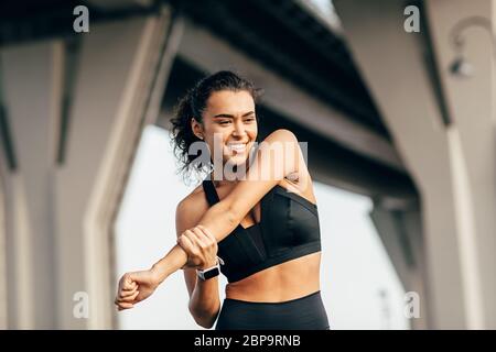 Woman athlete warming up her body before evening training under a highway. Smiling female stretching arm outdoors. Stock Photo