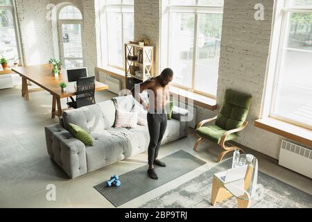 Young african-american man training at home during quarantine of coronavirus outbreak, doinc exercises of fitness, aerobic. Staying sportive during insulation. Wellness, movement concept. Stock Photo