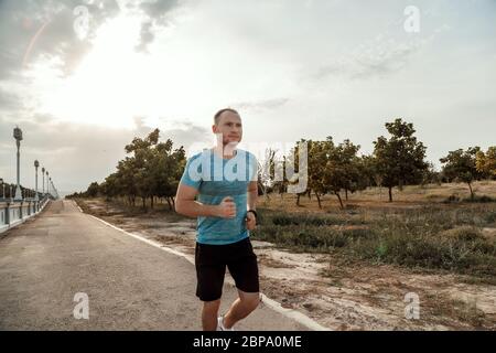 portrait of Caucasian guy in a blue t-shirt and black shorts who trains and runs on the asphalt track during sunset Stock Photo