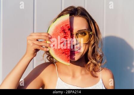 Funny curly woman in sunglasses hiding a half of her face with piece of watermelon, joking, smiling, standing against the wall, outdoors. Stock Photo
