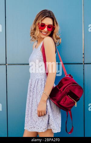 Attractive young woman in stylish red sunglasses, standing near the blue wall. Dressed in summer dress, with pink backpack. Outdoors. Stock Photo