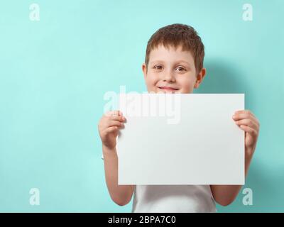 Four-year-old boy smiles and holds blank white paper sheet. Happy child on blue background with copy space for message, mock up Stock Photo