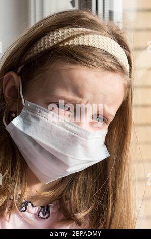 Small child with virus protection surgical face mask looking out the window, staying home for social distancing due to coronavirus Stock Photo