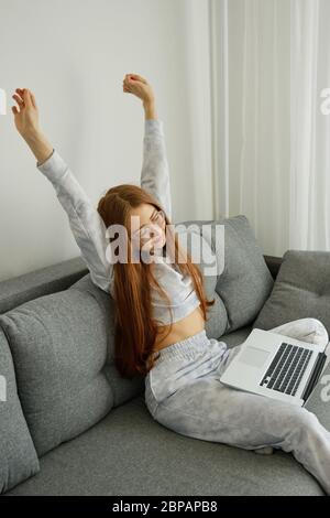Redhead girl in pajamas and glasses sits on a sofa with a laptop, stretching arms up Stock Photo