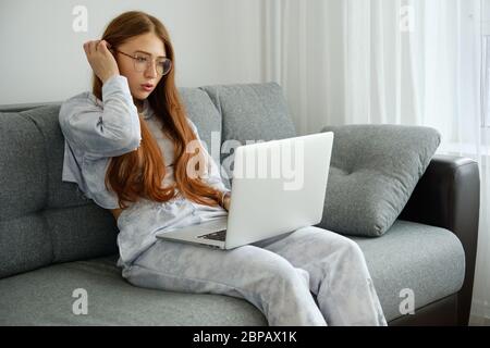 Redhead girl in pajamas and glasses sits on a sofa with a laptop, straightening her hair Stock Photo