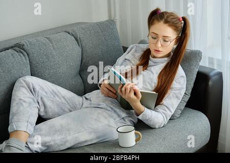 Red-haired girl with tails in pajamas lies on the sofa and reads a book Stock Photo