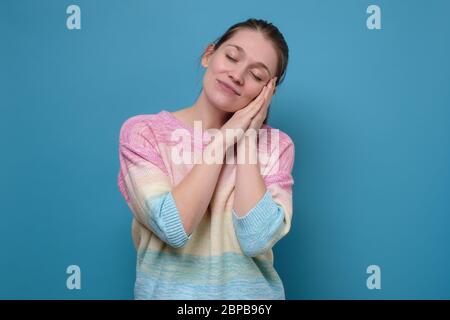 Sleepy caucasian young woman with closed eyes. Beautiful dreamy young woman holding hands near head on blue wall. Stock Photo